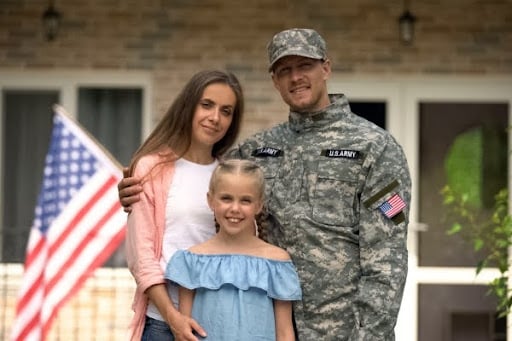 A smiling man in a U.S. Army uniform stands with a woman and a young girl, posing in front of a house with an American flag in the background.